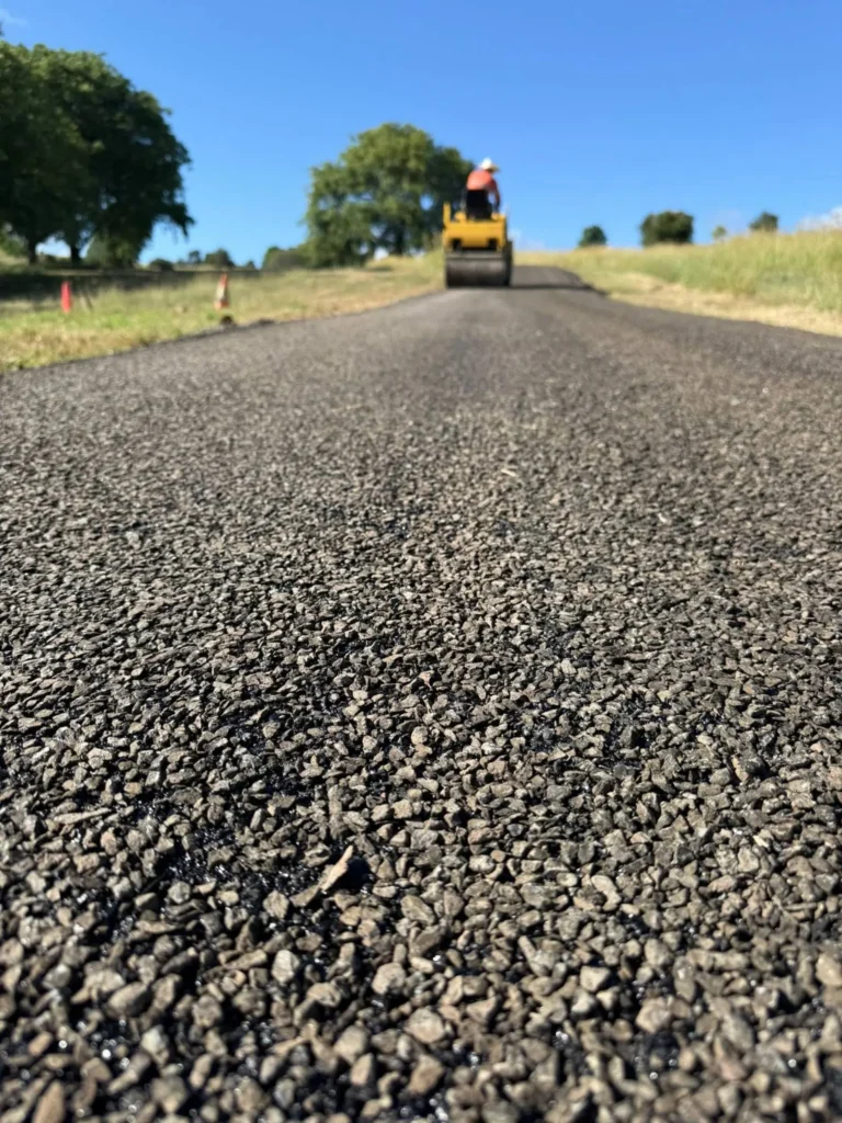 A freshly spray-sealed asphalt road with loose dark aggregate in the foreground, a road roller working in the distance.